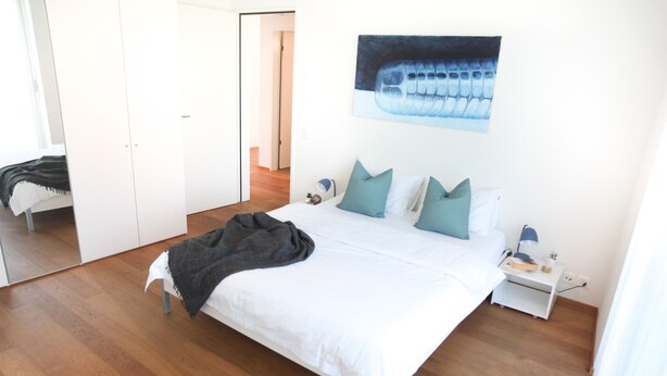 A bedroom with a bed, pillows, a nightstand, a lamp, and a painting on the wall.