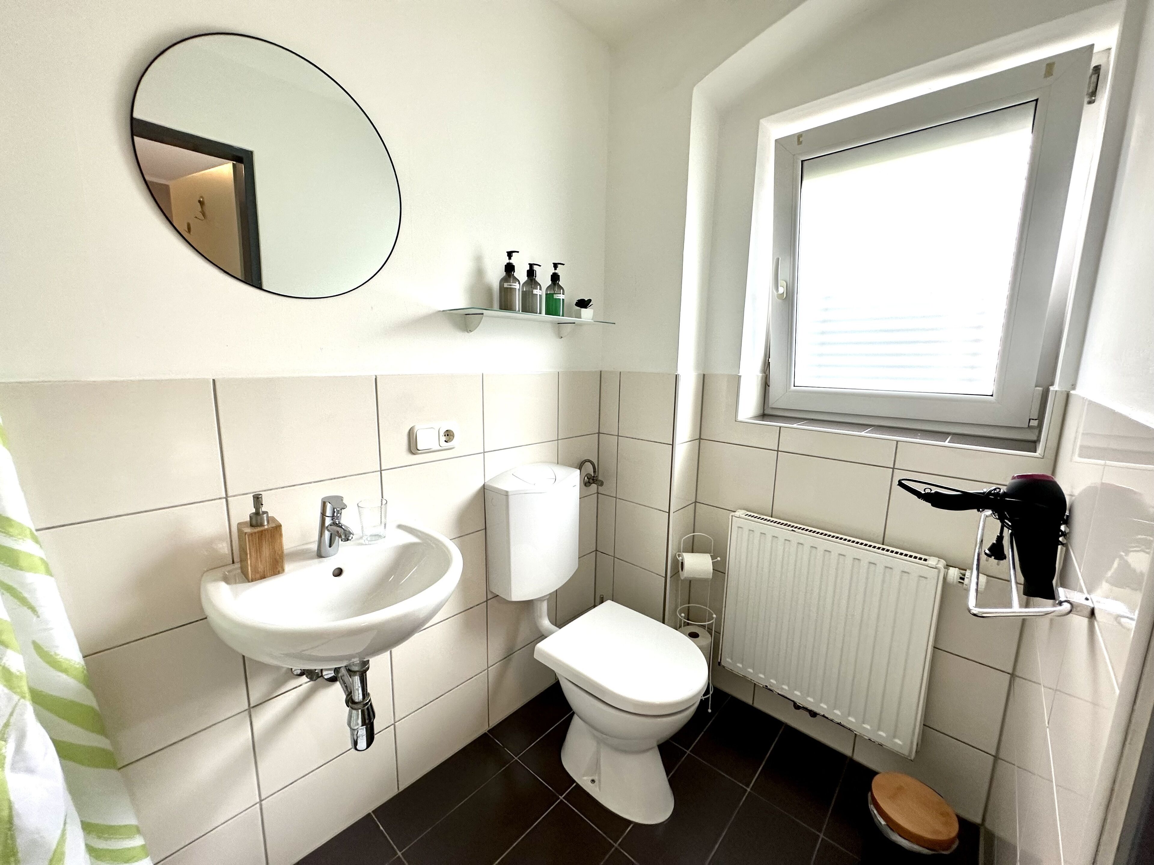 A bathroom with a white sink, toilet, and a mirror. There is a window above the toilet and a radiator below it.