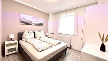 A modern bedroom with a bed, bedside tables, a radiator, a window with curtains, and a painting on the wall.