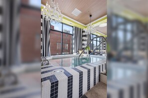 Uma área interna de piscina moderna com piso ladrilhado, um grande candelabro e vista para a cidade através de amplas janelas.