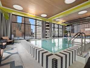 Piscina interior moderna com borda em azulejo, amplas janelas com vista para a cidade e área de estar com móveis de vime.