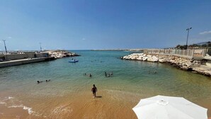 Een strand met helder water, een rotsachtige kade en iemand die in het water staat.