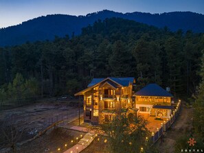A two-story lodge with a stone facade and a blue roof, surrounded by forested hills. - Zostel Pahalgam (Anantnag)