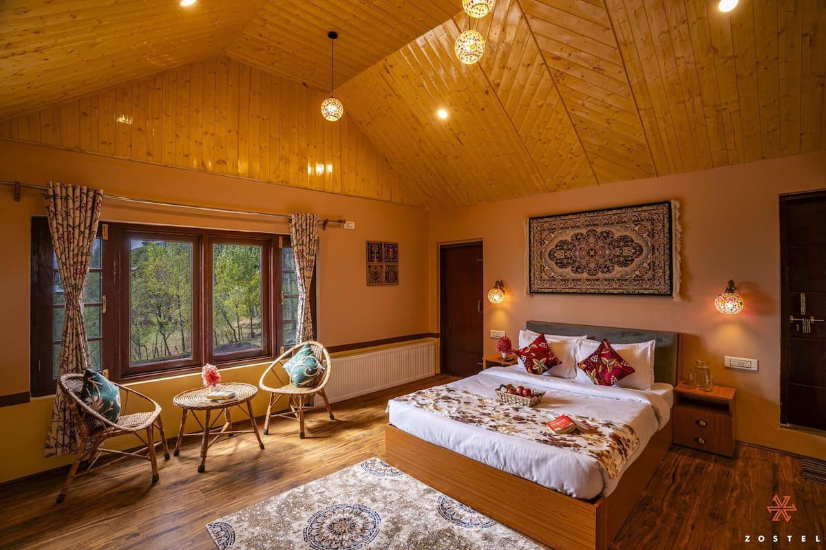 Um quarto aconchegante com teto de madeira, uma cama grande com roupa de cama estampada, duas cadeiras de vime, uma mesa pequena e vista para árvores pela janela.