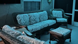 Un salon comprenant un canapé, deux fauteuils et une table basse. Un téléviseur à écran plat est fixé au mur, et il y a une fenêtre avec des stores.