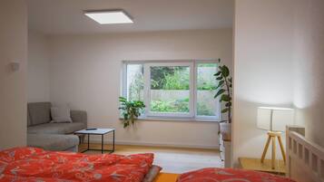 A room with a bed, a sofa, a small table, a lamp, and a window with a view of greenery.