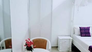 Une petite pièce blanche, avec un lit, une table de chevet, une chaise, une petite table sur laquelle est posé un vase de fleurs et une armoire.