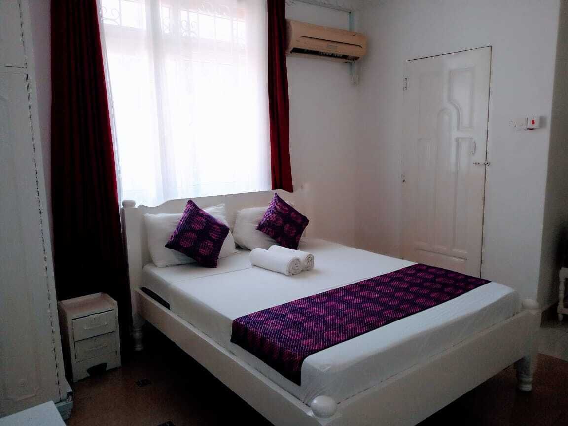Ein ordentlich bezogenes Bett mit Kissen und Decke, ein kleiner Nachttisch, ein Fenster mit Vorhängen und eine Tür.