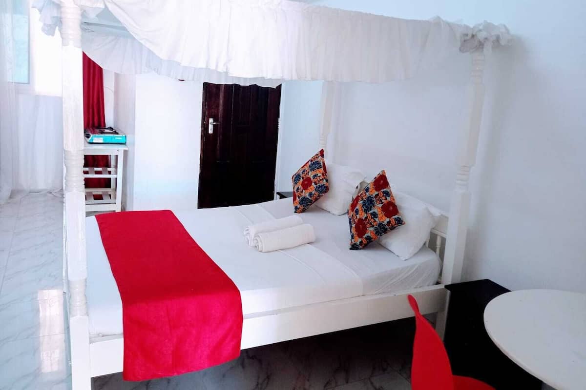 Una cama con dosel blanco con detalles rojos, una mesita y una silla roja en una habitación de hotel con suelo de mármol y una puerta oscura.