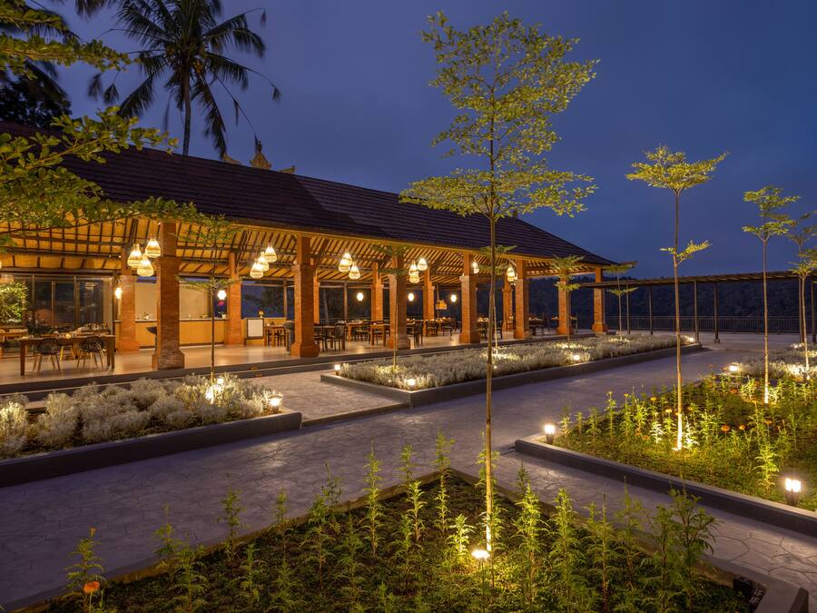 Un área de comedor al aire libre bien iluminada, con mesas y sillas, rodeada de jardines ajardinados y altos árboles.