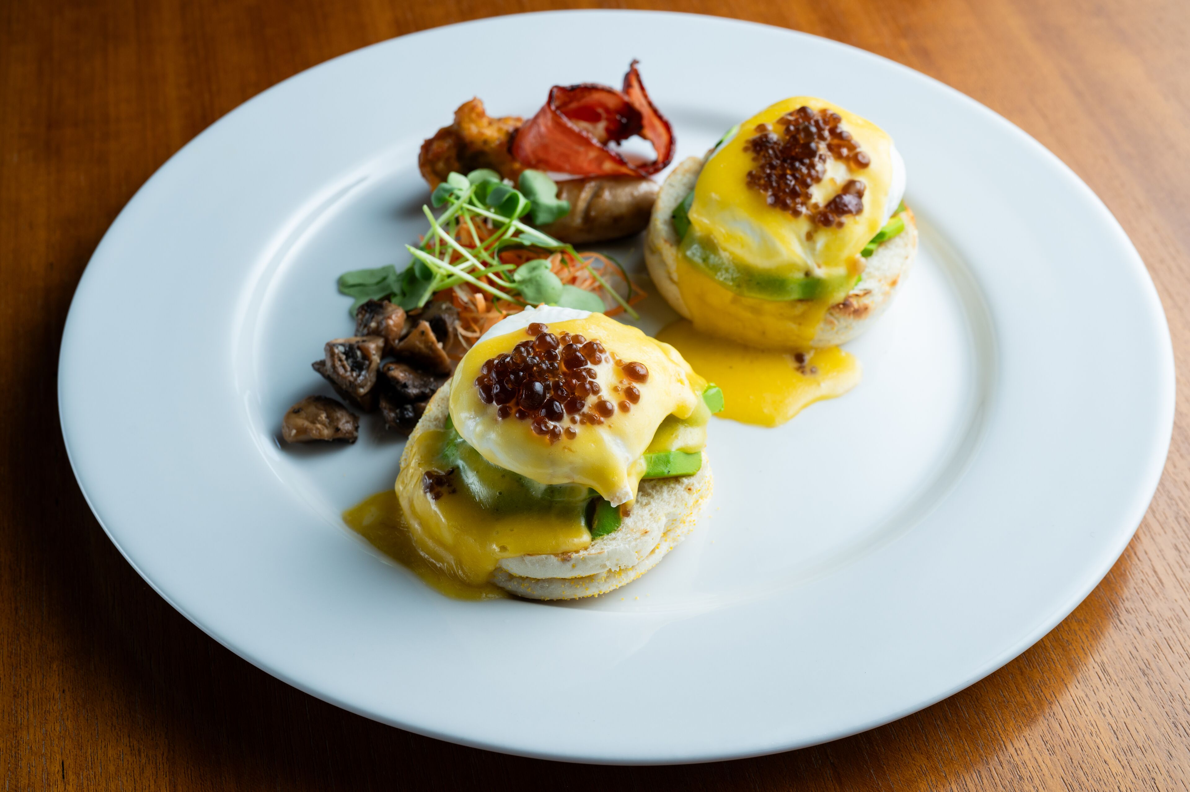 A white plate with two eggs benedict, bacon, mushrooms, and greens.