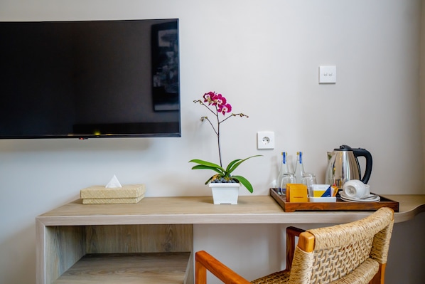 Habitación de hotel con televisor de pantalla plana montado en la pared, un escritorio de madera con una orquídea en maceta, una bandeja con botellas y un hervidor, y una silla de mimbre.