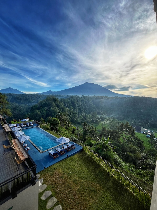 A pool with lounge chairs and umbrellas, surrounded by lush greenery and mountains in the background.