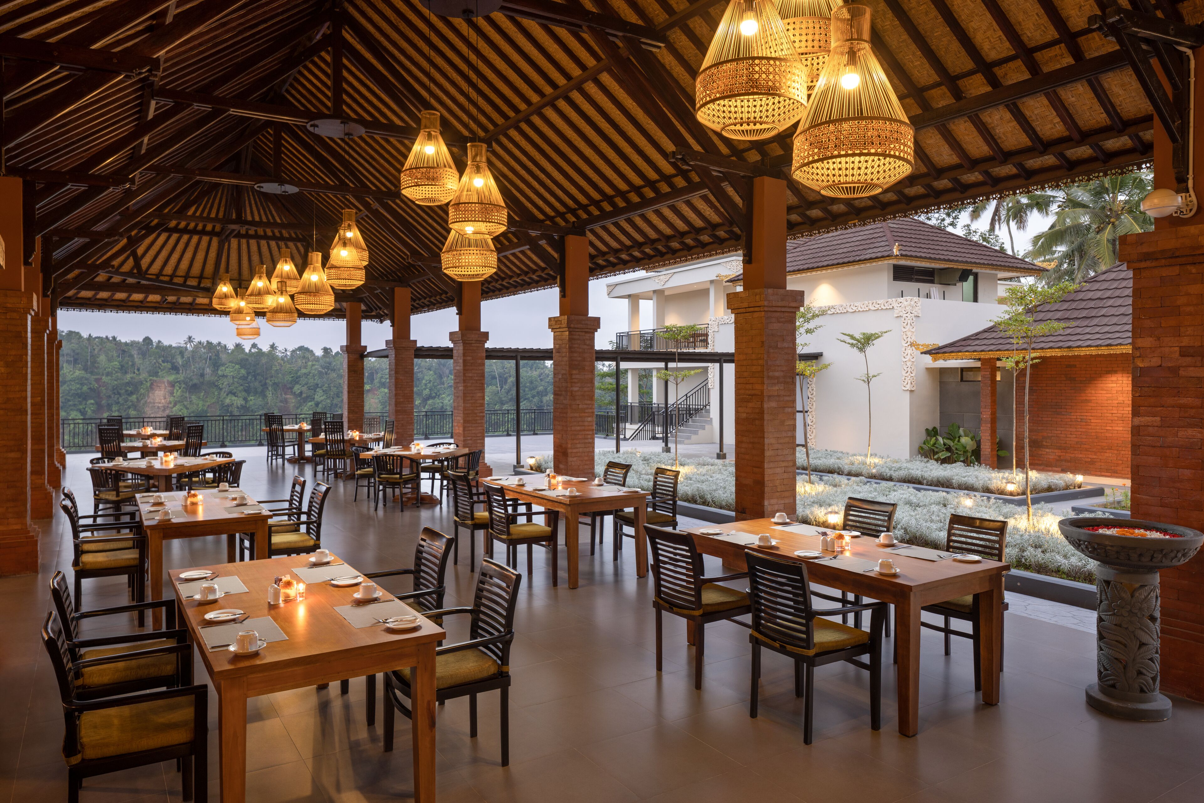 Un comedor interior con varias mesas y sillas, techo de paja y vistas a un edificio y follaje exterior.