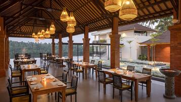 Un comedor interior con varias mesas y sillas, techo de paja y vistas a un edificio y follaje exterior.