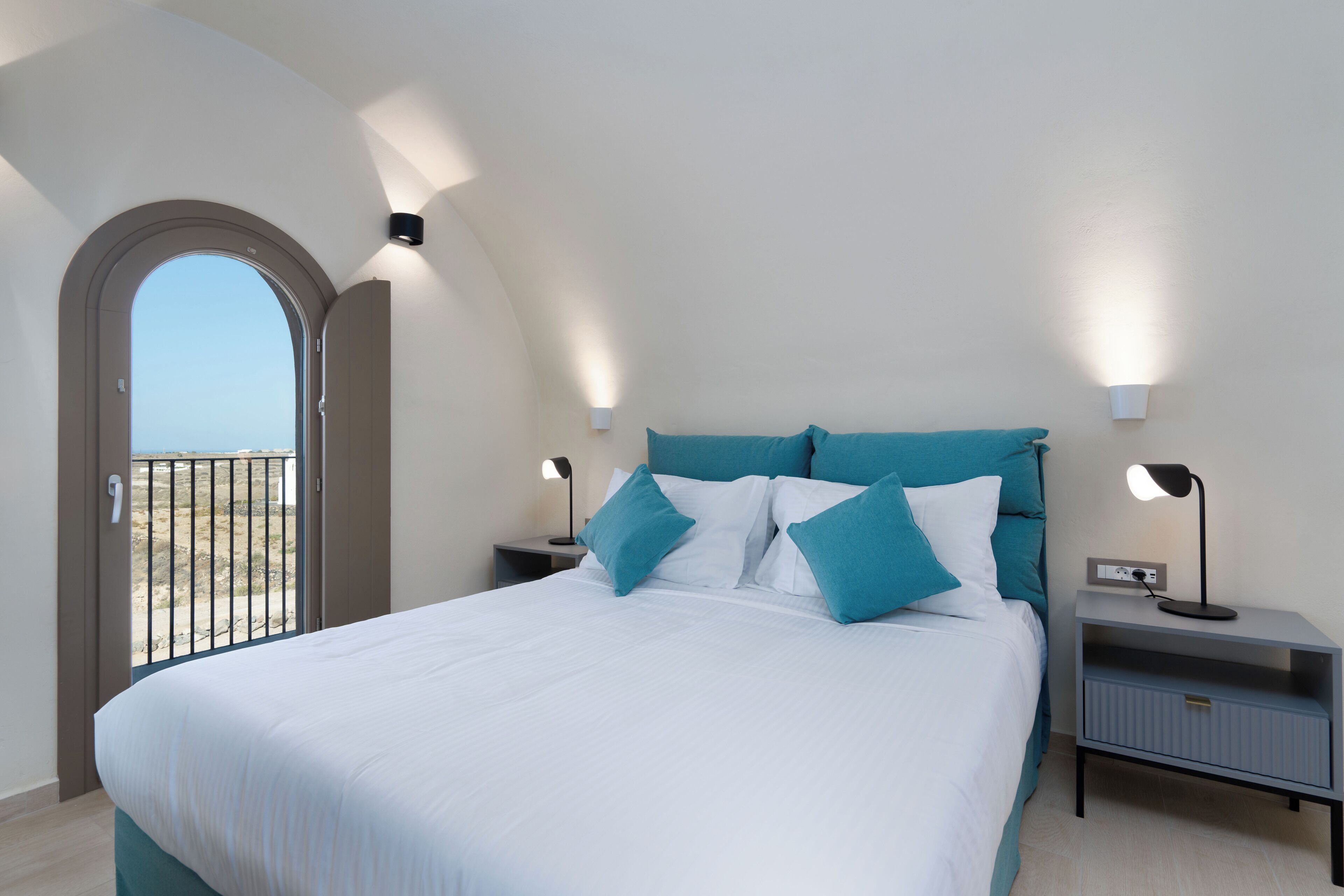 A bedroom with a large bed, two bedside tables with lamps, a view through an arched doorway, and a balcony.