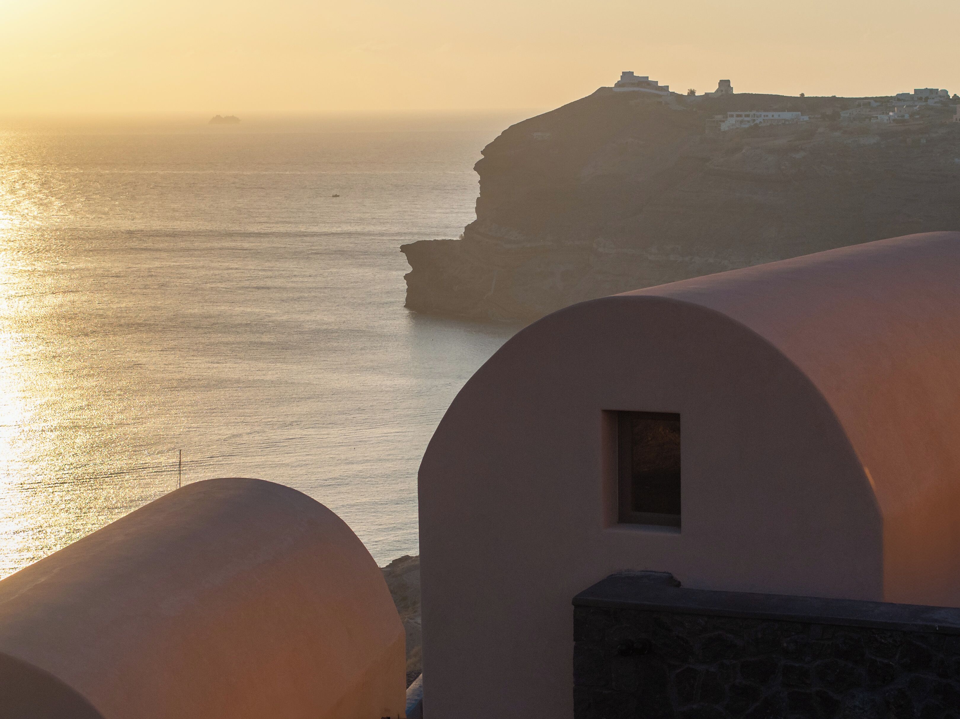 A coastal view with a building featuring a small window and a stone base, overlooking the sea and a cliff with structures on top.