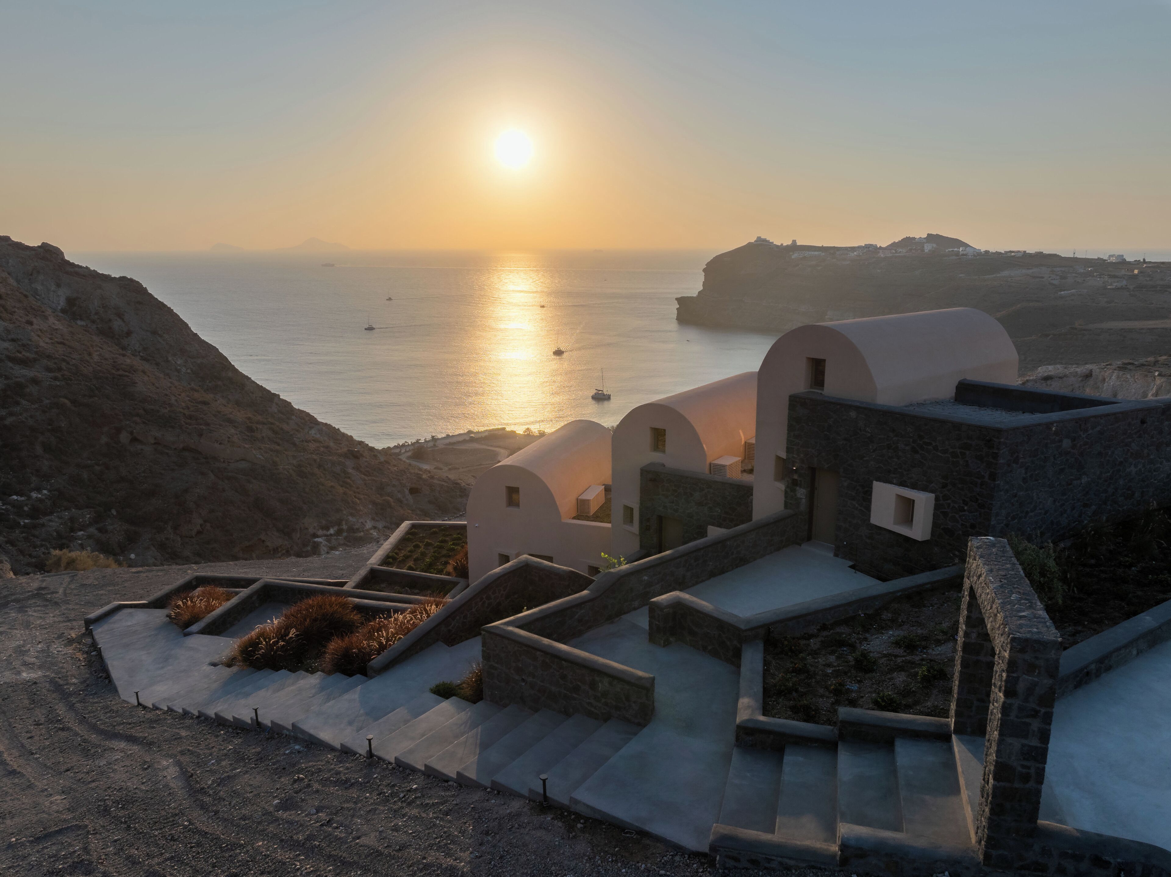 A coastal building with a stone facade, a staircase leading to a terrace, and a view of the sea and sunset.