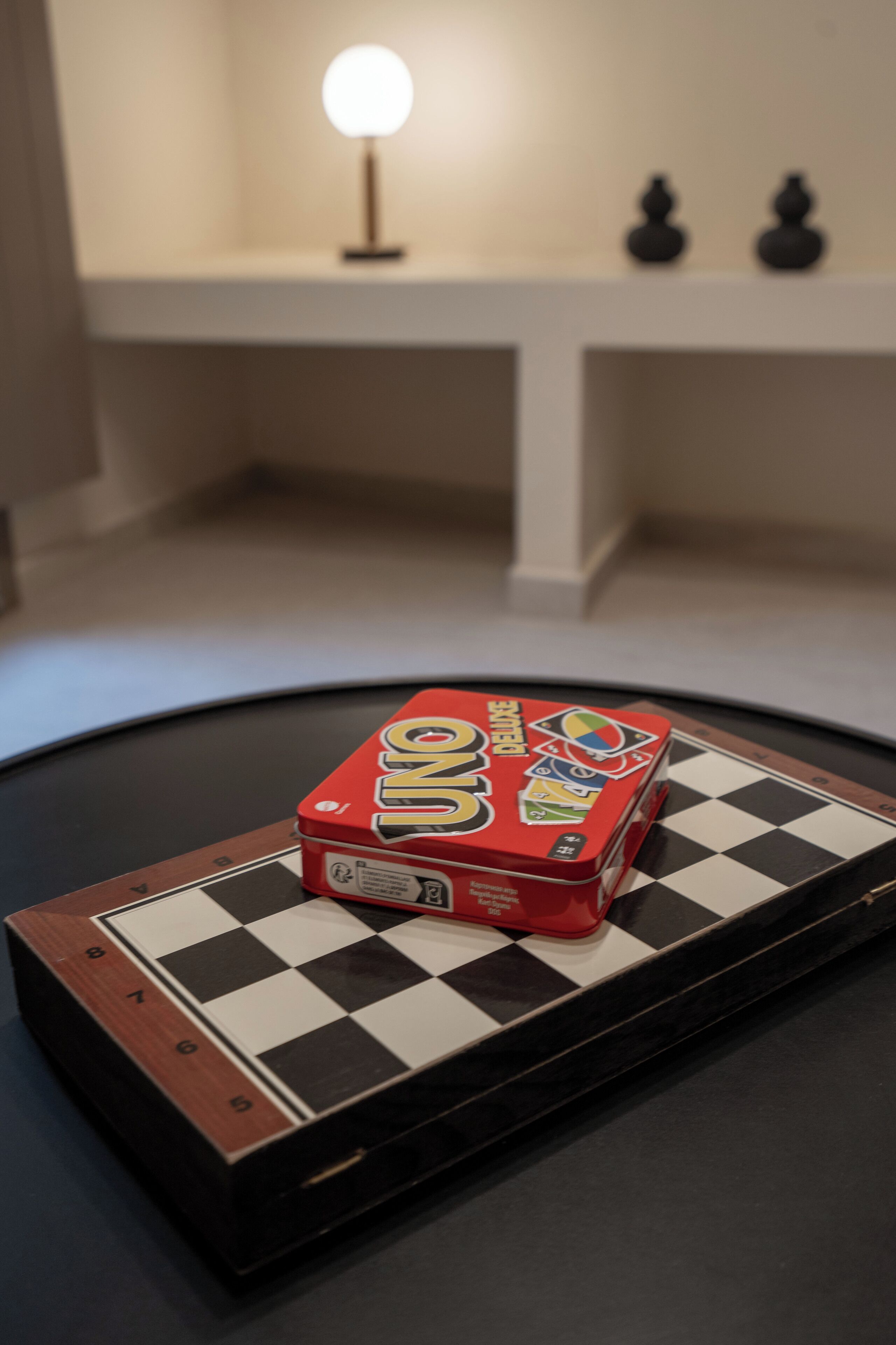 A chessboard with a box of UNO cards on top, a lamp, and two figurines on a white shelf in the background.