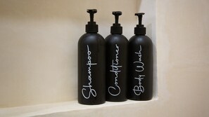 Três frascos pretos com bombas dosadoras, identificados como 'Shampoo', 'Condicionador' e 'Gel de Banho', em uma prateleira.
