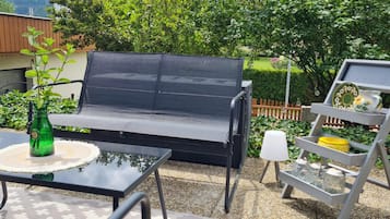 Un canapé noir pour l’extérieur, agrémenté d’un coussin, une petite table ronde avec une bouteille verte et une lampe blanche.