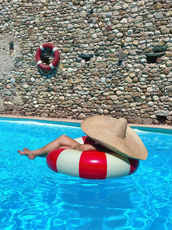 A person floating in a pool with a red and white inflatable ring, wearing a straw hat, and a life preserver hanging on a stone wall. - Les Cailloux Dorés (Massillargues-Attuech)