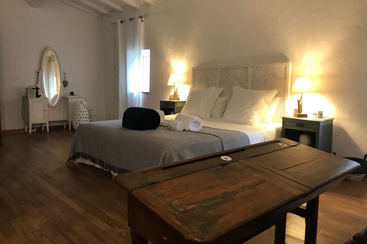 Una habitación de hotel con cama, mesitas de noche con lámparas, un banco de madera, un tocador con espejo y una ventana con cortinas.