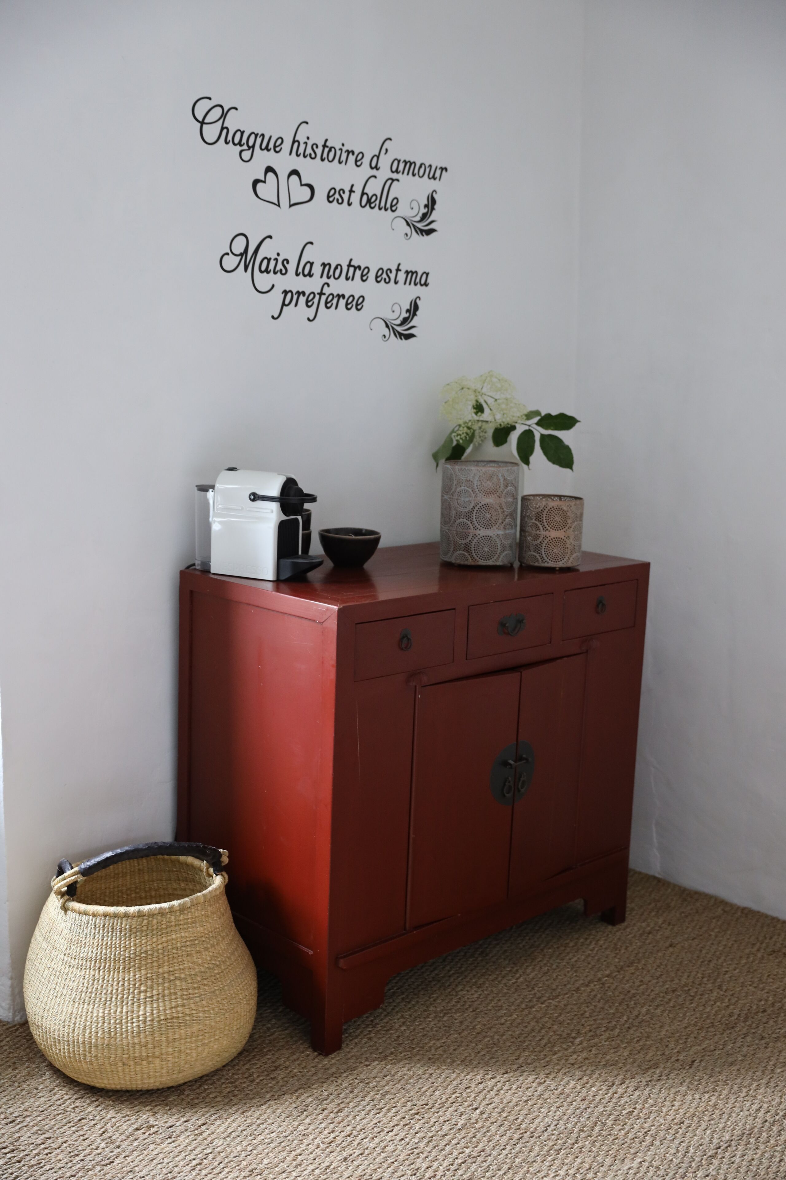 A red cabinet with a white appliance on top, a decorative wall decal, and a woven basket beside it.