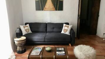 A living room with a black sofa, two matching pillows, a woven stool, a white fluffy ottoman, and a coffee table with suitcases and magazines.