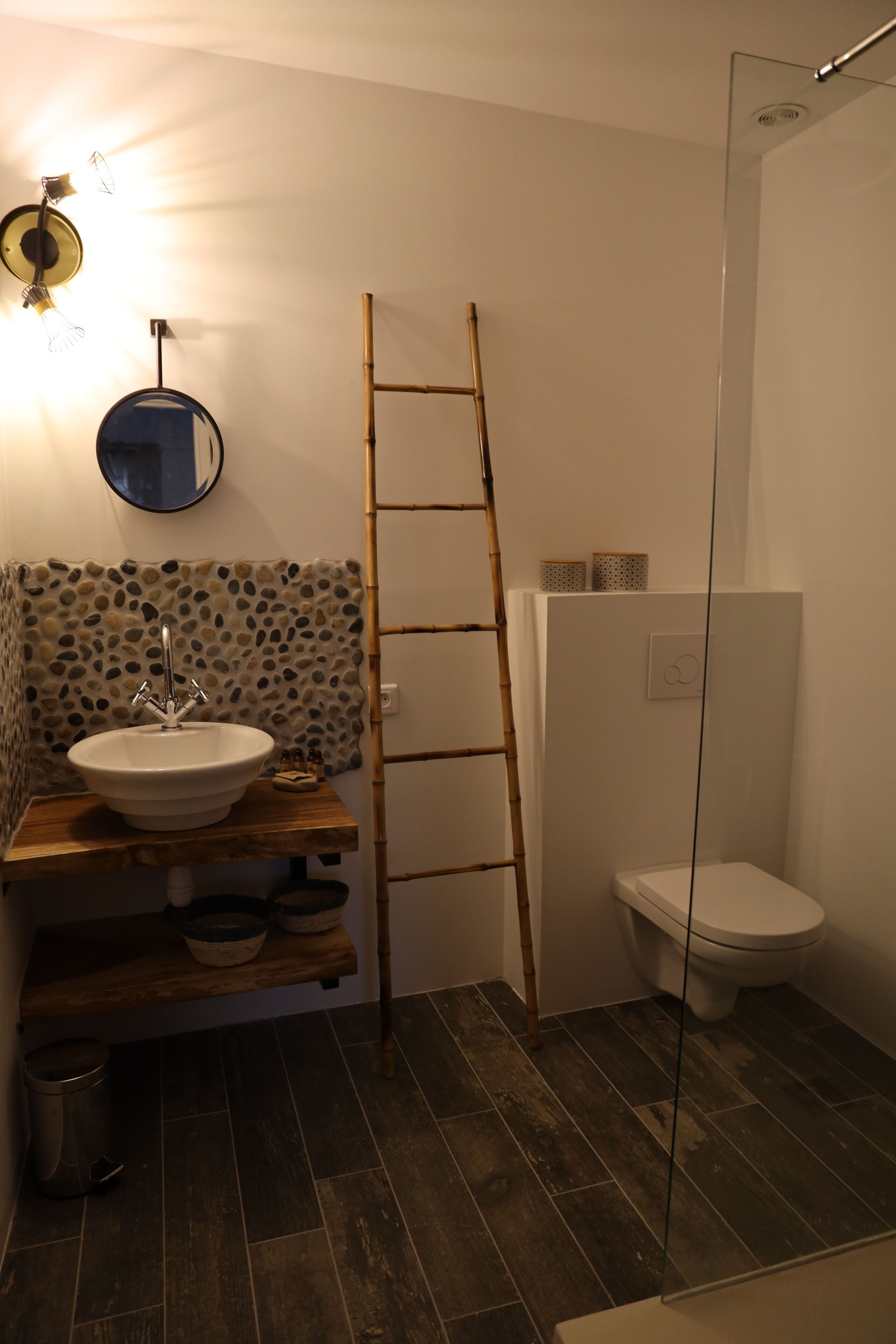A bathroom with a wooden shelf holding bowls, a white sink, a bamboo ladder, a toilet, and a shower area.