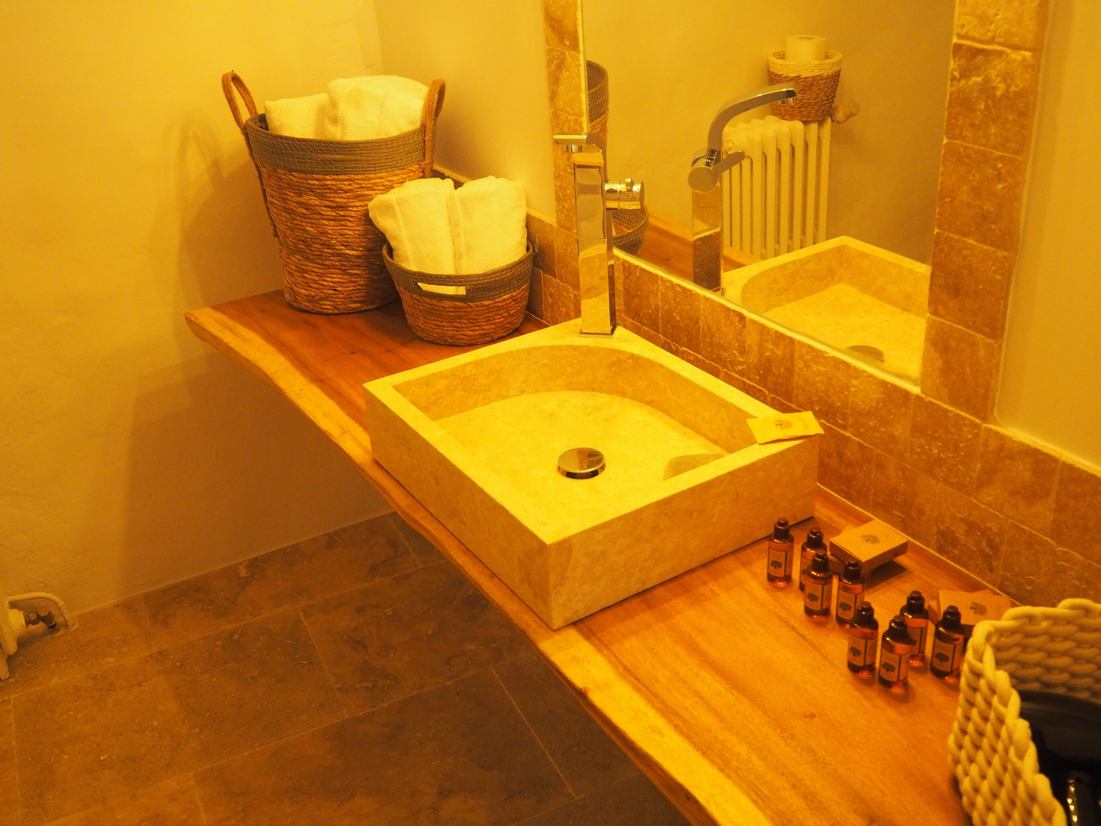 Une salle de bain avec un meuble-lavabo en bois, un grand lavabo rectangulaire et un miroir. On peut voir deux paniers en osier posés sur le meuble-lavabo, et un flacon de savon liquide est visible sur le comptoir.