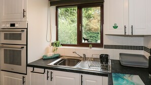 A kitchen with white cabinets, a black countertop, a window with a view of greenery, and a first aid box on the wall. - Harvey's Holiday House (Hunstanton)