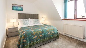 A bedroom with a bed, two bedside tables, a nightstand, a window with curtains, a radiator, and a ceiling light fixture. - Harvey's Holiday House (Hunstanton)