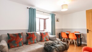 A living room with a sofa, a furry ottoman, and a dining area with orange chairs. - Harvey's Holiday House (Hunstanton)