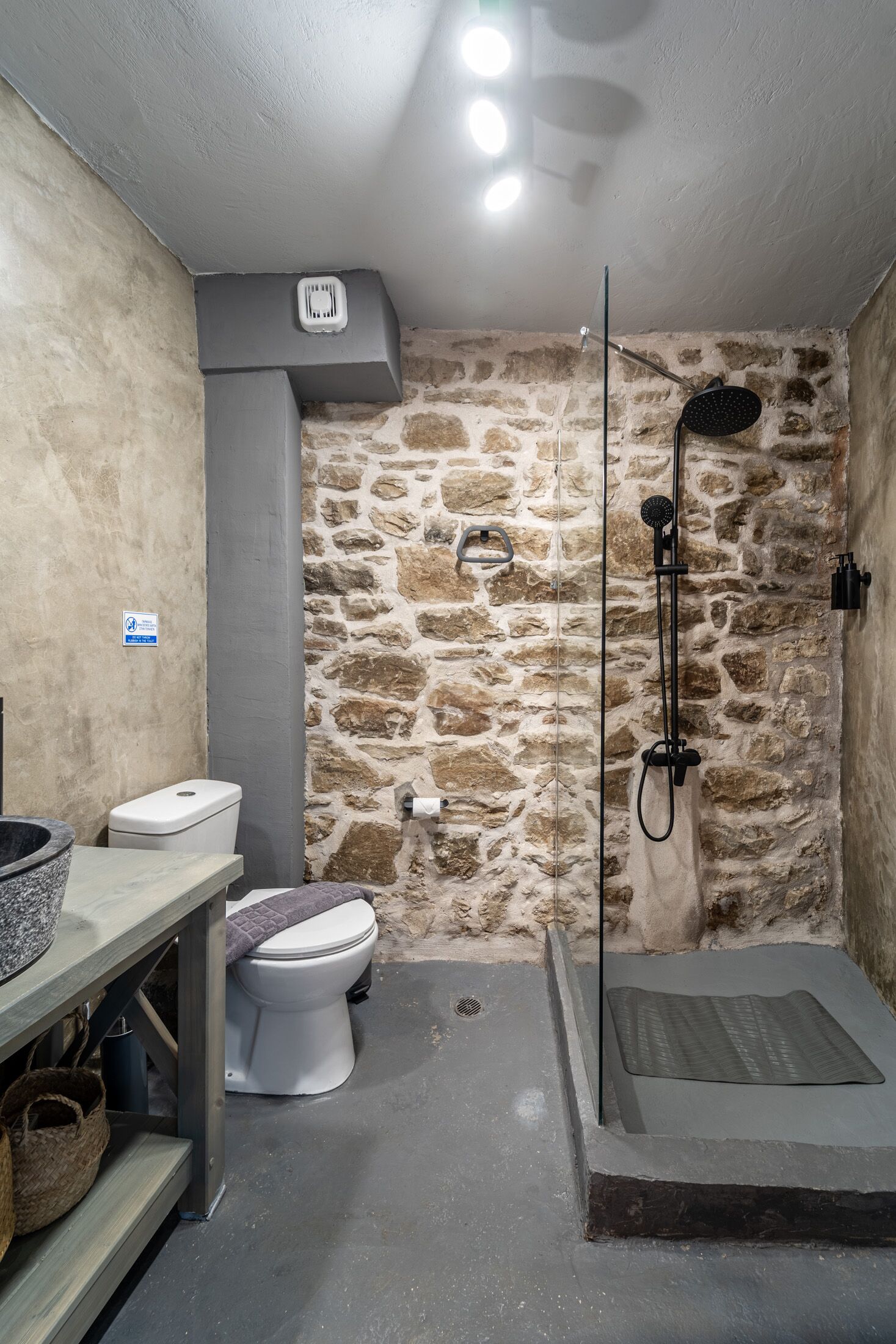 Un baño con pared de piedra, inodoro, ducha con puerta de vidrio y lavabo con encimera de granito.