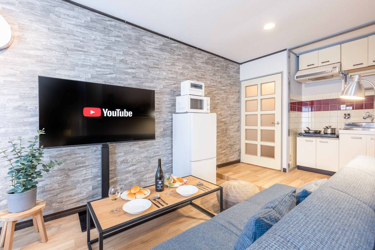 A modern living room with a flat-screen TV, a dining table set with plates and a bottle, a sofa, and a kitchen area with cabinets and a microwave.