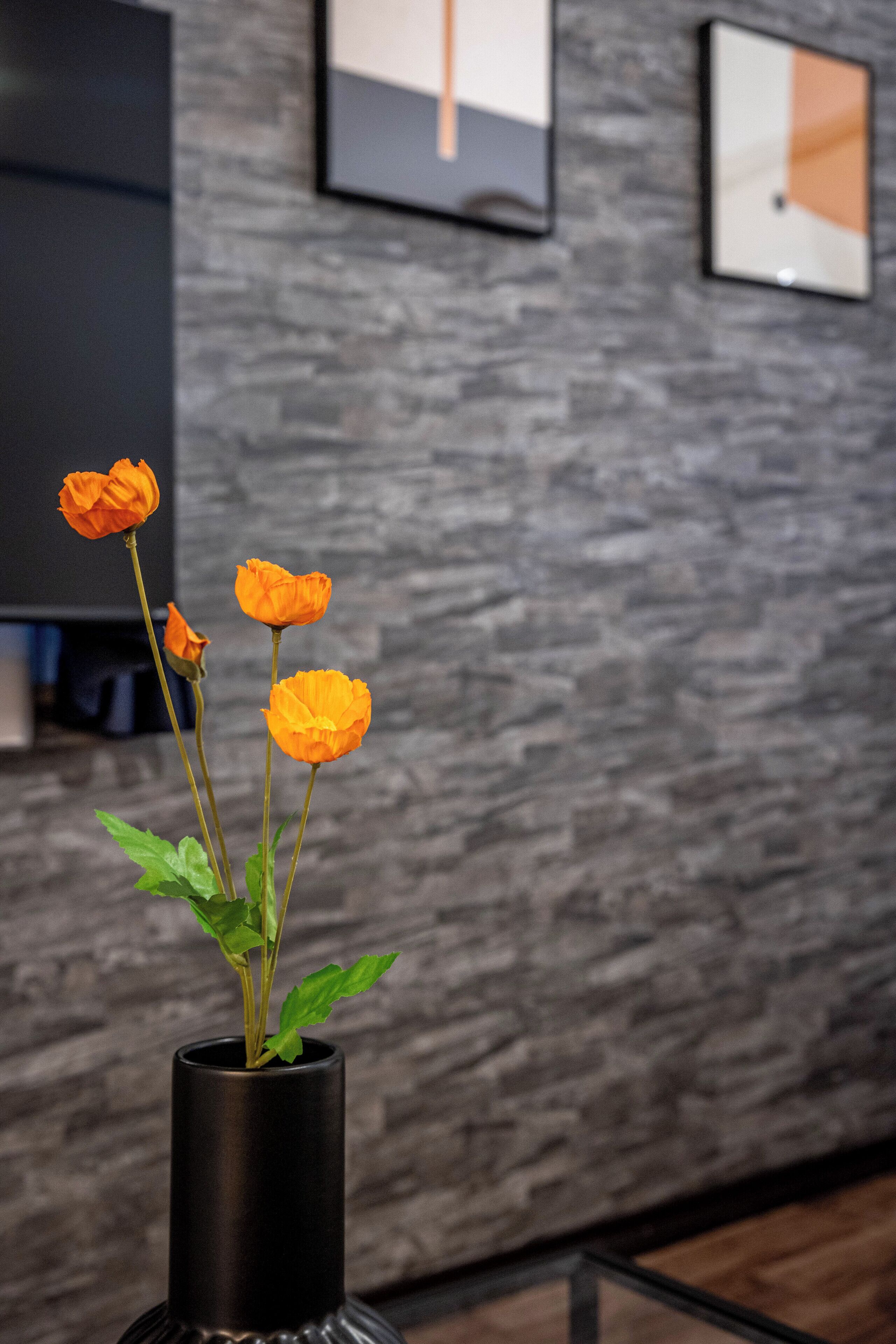 Un jarrón negro con flores naranjas sobre una mesa de vidrio frente a una pared gris con textura y cuadros enmarcados.
