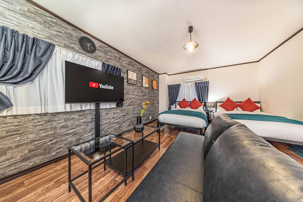 A modern living room with a flat-screen TV, a sofa, a coffee table, and a bed with red pillows.