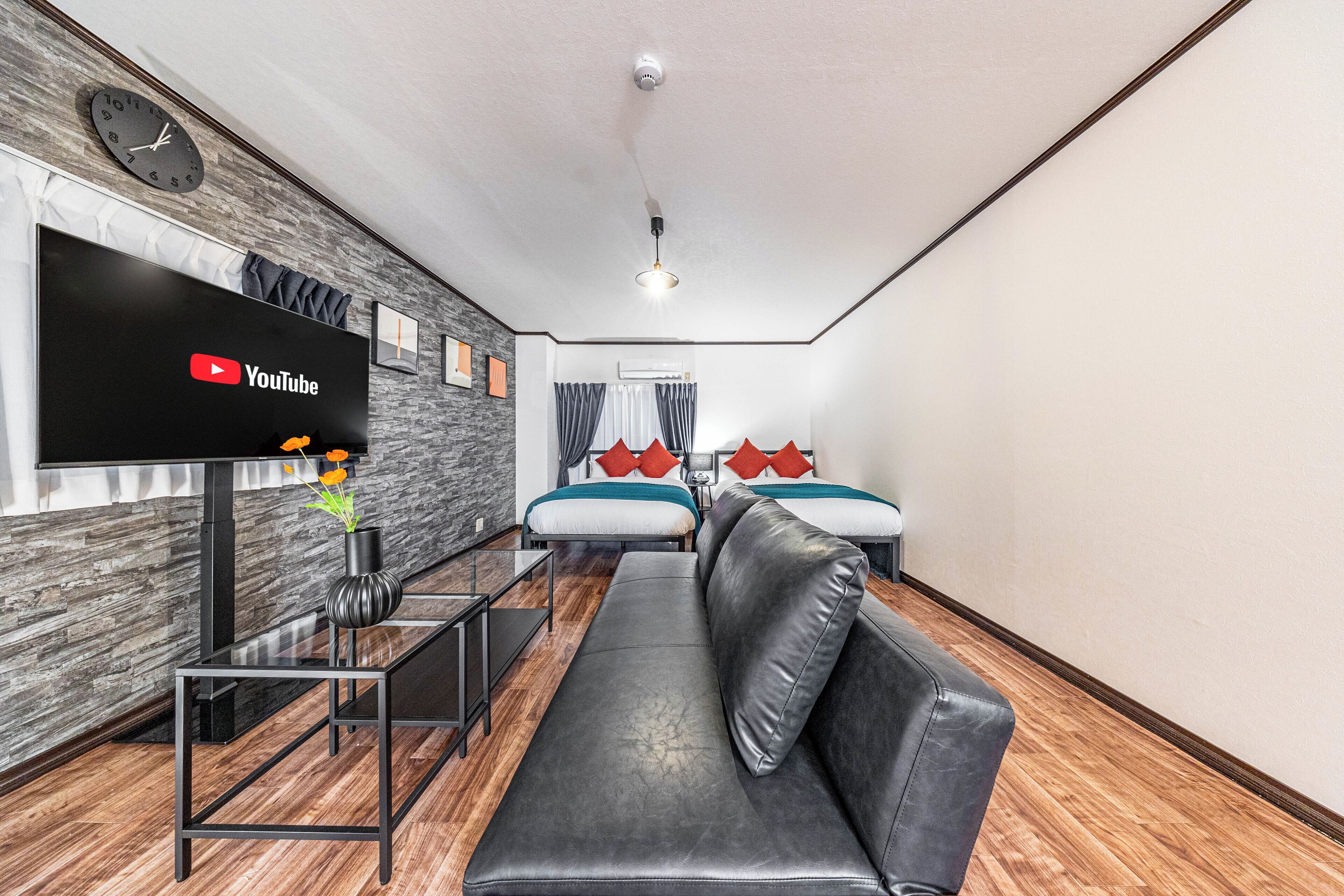 A modern hotel room with a flat-screen TV, a sofa, two beds, a wooden floor, and a clock on the wall.