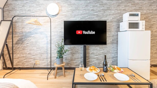 Una sala moderna con un televisor de pantalla plana montado en una pared con textura de piedra, una mesa de comedor dispuesta con platos, cubiertos y una botella de vino, y un refrigerador blanco a la derecha.