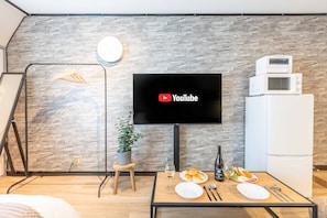 Ein modernes Wohnzimmer mit einem Flachbildfernseher an einer Wand mit Steinoptik, einem Esstisch gedeckt mit Tellern, Besteck und einer Weinflasche, sowie einem weißen Kühlschrank auf der rechten Seite.