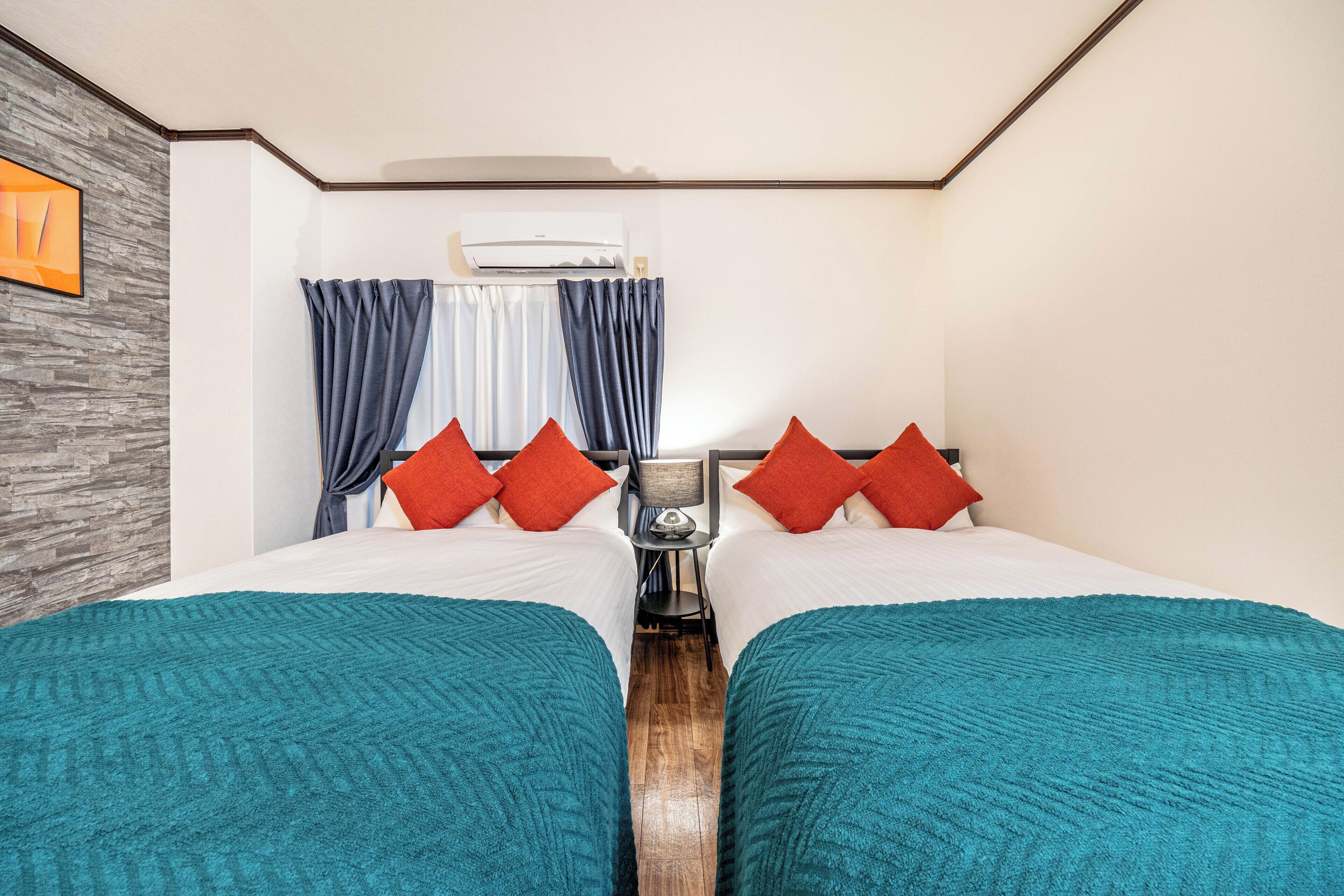 A hotel room with two beds, teal blankets, red pillows, a nightstand, and a wall-mounted air conditioner.