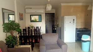 Un salon comprenant un canapé gris, un canapé marron, une table à manger avec des chaises, une plante en pot et un coin cuisine avec une cuisinière, un réfrigérateur et une poubelle.