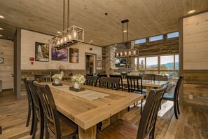 A spacious dining area with a large wooden table and chairs, surrounded by stone walls and large windows offering a view of the outdoors.