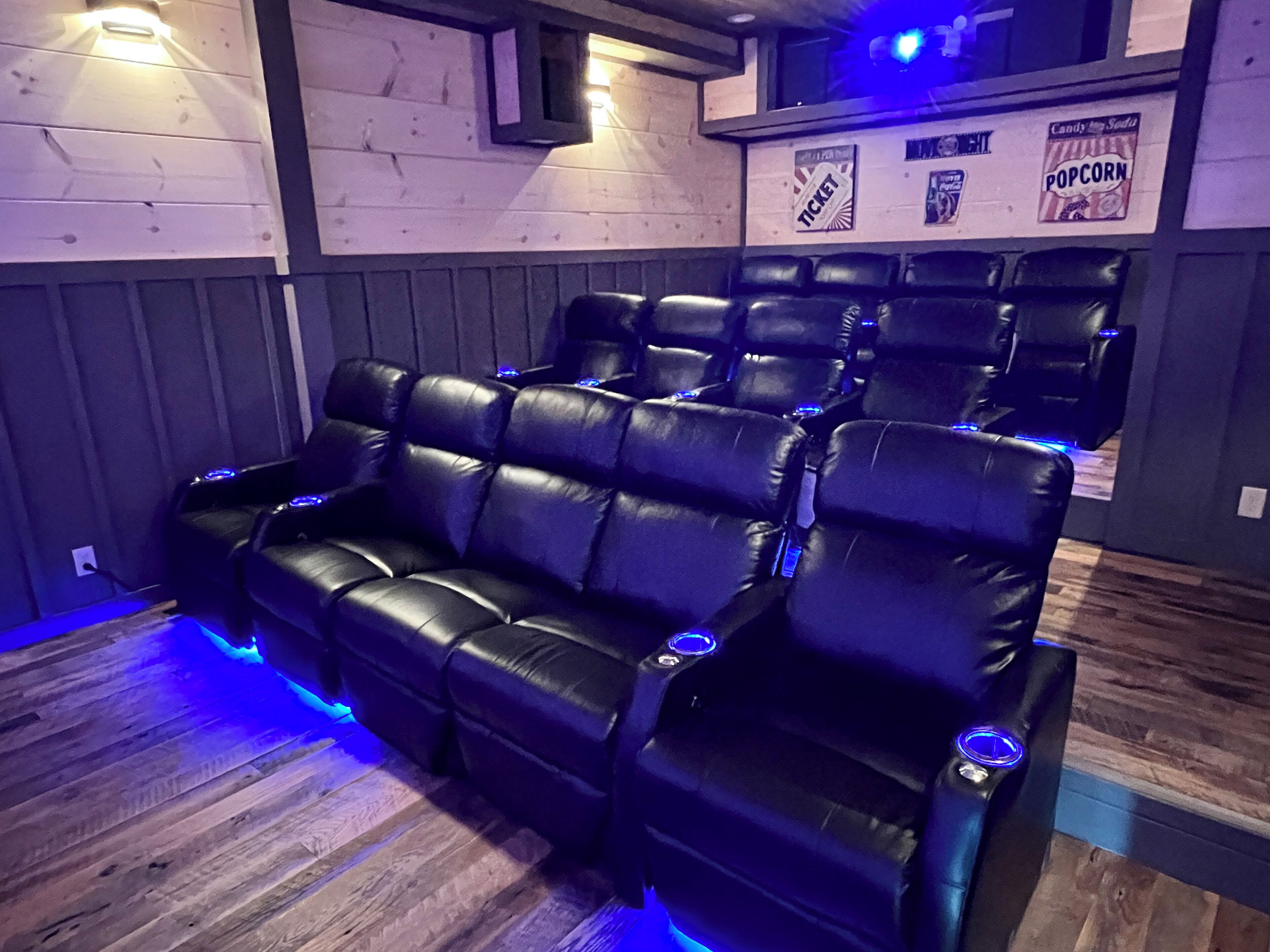 A row of black leather recliner seats with blue ambient lighting, wooden panel walls, and a popcorn sign on the wall.