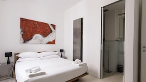 Une chambre à coucher moderne avec un grand lit, un tableau, un miroir et une salle de bain visible à travers une porte ouverte.