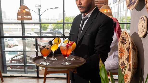 A waiter carrying a tray with three drinks in a modern interior with large windows and woven wall decorations. - Laten Suites Al Salim Plaza (Jeddah)