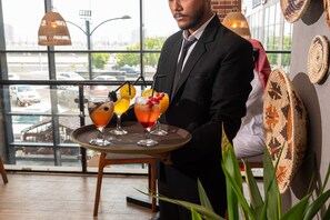 Un serveur portant un plateau avec trois boissons, dans un intérieur moderne doté de grandes fenêtres et de décorations murales en fibres tressées.