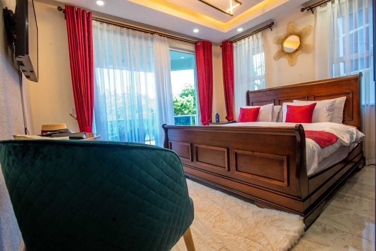 Quarto com cama de madeira, cortinas vermelhas e brancas, uma cadeira verde e vista para o exterior com vegetação.