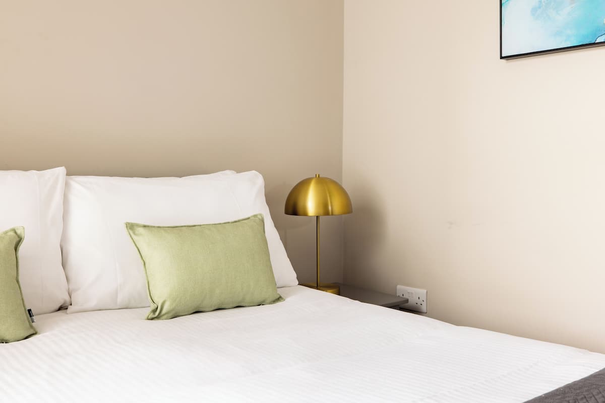 Een net opgemaakt bed met witte lakens en twee groene kussens, een goudkleurige vloerlamp en een stopcontact aan de muur.